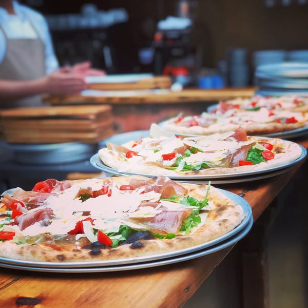 Several freshly baked pizzas with prosciutto, arugula, cherry tomatoes, and shaved cheese on a wooden counter.