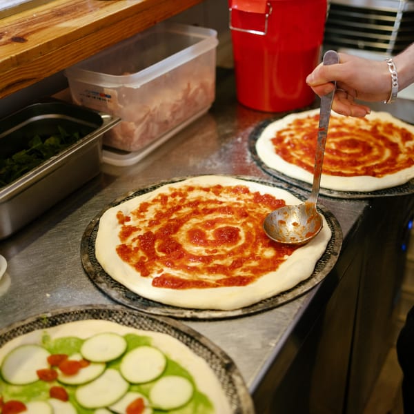 A chef spreads tomato sauce on pizza dough, with other pizzas and ingredients nearby.